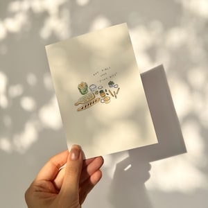 Può includere: Una carta color crema con un'illustrazione disegnata a mano di vari alimenti, tra cui pane, salse e guarnizioni. Il testo "Hot Girls Love - Picky Bites" è scritto in alto. La carta è tenuta da una mano.