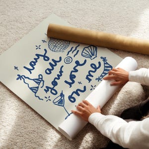 May include: A person is rolling up a white poster with blue text that says "lay your worries on me". The poster has a blue and white party hat, a seashell, and a disco ball drawn on it.