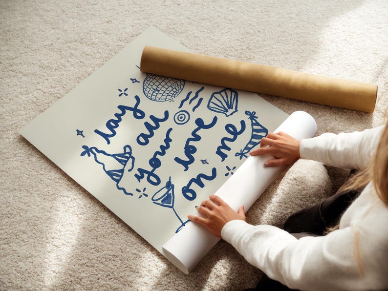 May include: A person is rolling up a white poster with blue text that says "lay your worries on me". The poster has a blue and white party hat, a seashell, and a disco ball drawn on it.
