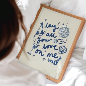 May include: A framed print with a white background and blue text that reads "lay all your love on me". The text is surrounded by illustrations of a cocktail glass, a disco ball, a seashell, and a party hat.