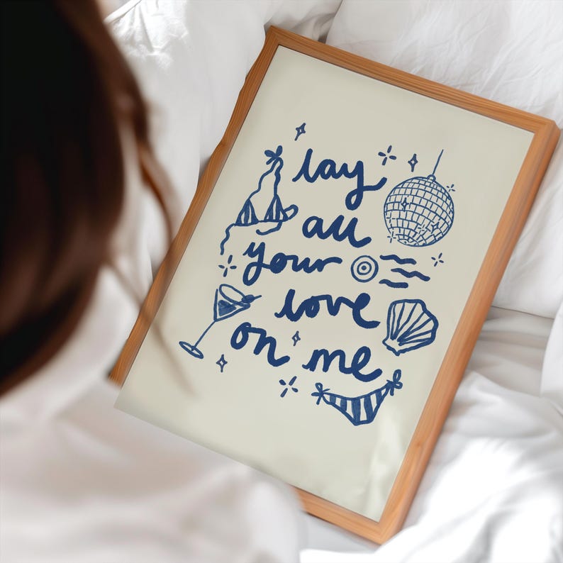 May include: A framed print with a white background and blue text that reads "lay all your love on me". The text is surrounded by illustrations of a cocktail glass, a disco ball, a seashell, and a party hat.