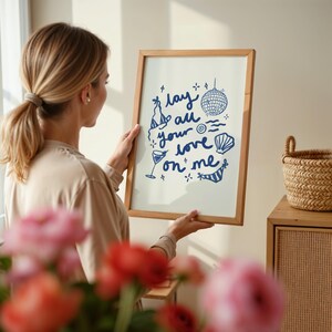 May include: A framed print with a blue and white design that says "Lay all your love on me". The print is surrounded by a wooden frame and is hanging on a wall.