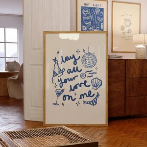 May include: A framed print with a blue and white illustration of a disco ball, a cocktail glass, and a seashell. The text "Lay all your love on me" is written in a whimsical font.