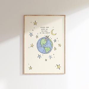 May include: A framed art print featuring a whimsical illustration. The artwork depicts a blue and green globe with two figures standing on top, surrounded by stars and a crescent moon. Text reads "Young and Dumb on the Edge of the World."