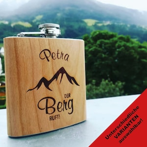 Könnte beinhalten: Eine Holzflasche mit silbernem Verschluss. Die Flasche ist mit dem Namen "Petra" und den Worten "Der Berg Ruft!" graviert. Die Flasche steht vor einem verschwommenen Hintergrund eines Berges.