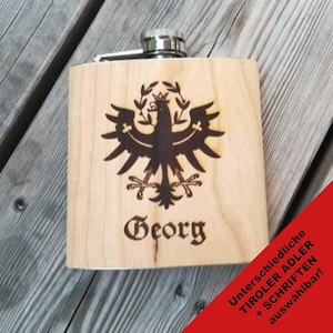 Könnte beinhalten: Eine Holzflasche mit einem Metallverschluss, die mit einem schwarz-weißen Adlermotiv und dem Namen "Georg" auf der Vorderseite verziert ist. Die Flasche liegt auf einer Holzoberfläche. Der Text "Unterschiedliche Tiroler Adler + Schriftarten auswählbar!" ist in der unteren rechten Ecke sichtbar.
