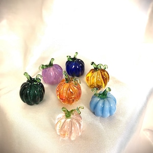 Könnte beinhalten: Sieben kleine Glaskürbisse in verschiedenen Farben, darunter grün, lila, blau, orange, gelb, hellblau und rosa. Die Kürbisse sind alle etwa 5 cm hoch und haben eine realistische Kürbisform.
