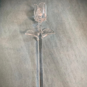 Puede incluir: Una escultura de rosa de cristal transparente con un tallo largo y dos hojas. La rosa tiene un diseño detallado de pétalos y está hecha de vidrio transparente. La escultura es un objeto decorativo.