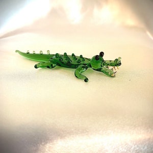 May include: A green glass alligator figurine with black eyes and a detailed textured body. The alligator is posed with its mouth open and its teeth visible.