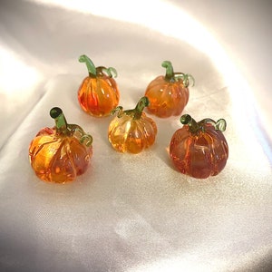 May include: Five miniature glass pumpkins in shades of orange and amber, each with a green stem. The pumpkins are arranged on a white, textured surface, creating a seasonal or fall theme.