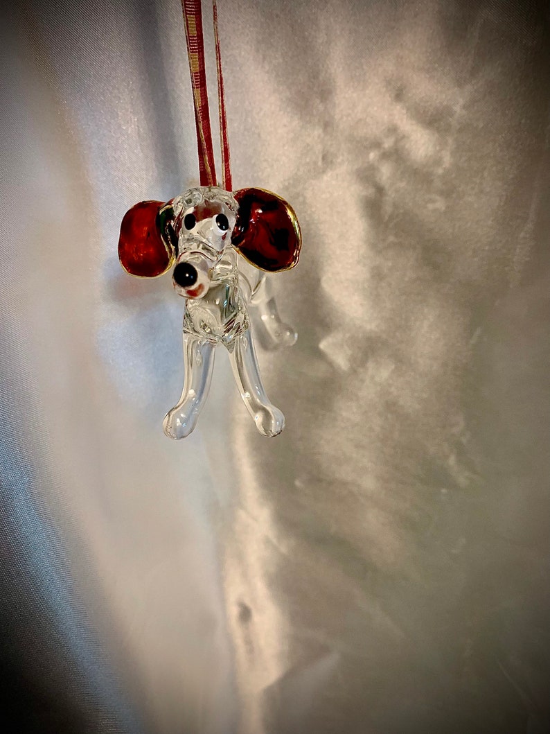 May include: A clear glass ornament shaped like a dog with red and gold ears. The ornament is hanging from a red and white ribbon.