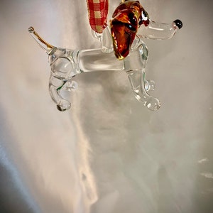 May include: A glass dachshund ornament with a brown and amber head and black eyes. The ornament is hanging from a red and white gingham ribbon.