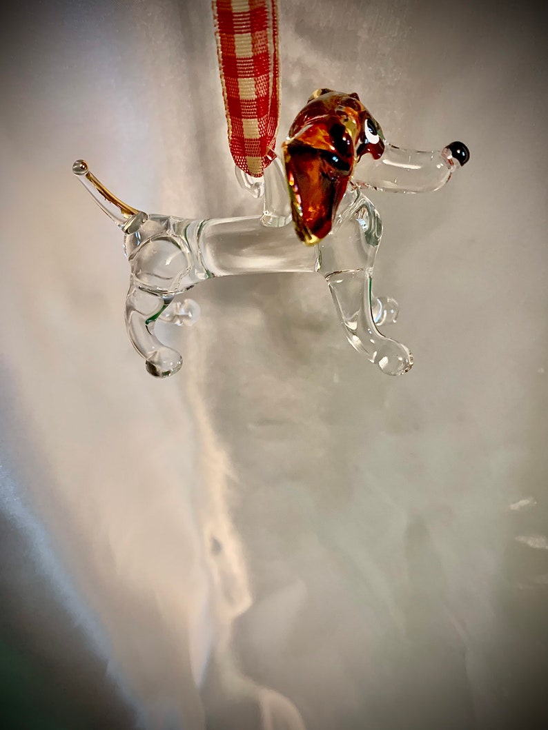 May include: A glass dachshund ornament with a brown and amber head and black eyes. The ornament is hanging from a red and white gingham ribbon.