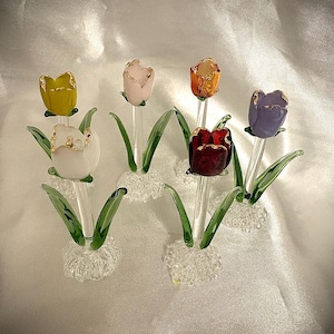 Könnte beinhalten: Sechs gläserne Tulpen in verschiedenen Farben, darunter Gelb, Weiß, Rosa, Orange, Rot und Lila, mit goldenen Verzierungen. Jede Tulpe steht auf einem klaren Glasstiel mit grünen Glasblättern und einem weißen Glasfuß.