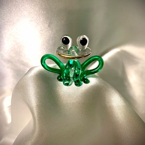 以下が含まれることがあります： 黒い目をした緑色のガラス製のカエルの置物。口の周りに金色の縁取りがあります。カエルは白いサテン生地の上に座っています。