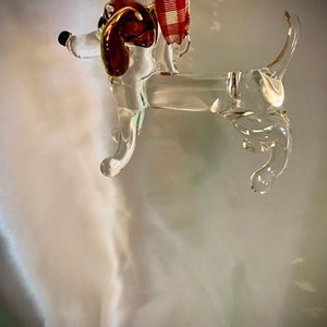 May include: A clear glass ornament shaped like a dachshund dog with red glass ears. The ornament has a red and white checkered ribbon attached to the top.