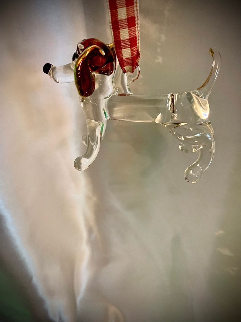 May include: A clear glass ornament shaped like a dachshund dog with red glass ears. The ornament has a red and white checkered ribbon attached to the top.