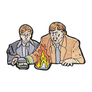 May include: Embroidered patch featuring a scene with two men, one in a gray suit and the other in a tan blazer. The man in the blazer has a surprised expression and a flame is in front of him. The patch has a black border.