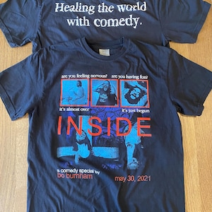 Peut inclure: T-shirt noir avec un graphique bleu et rouge du spectacle "Inside" du comédien Bo Burnham. Le texte "Healing the world with comedy" est imprimé au dos du t-shirt.