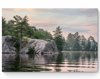 May include: A rocky island with trees and a calm lake in the foreground. The sky is a soft pink and blue with clouds. The water reflects the sky and trees.