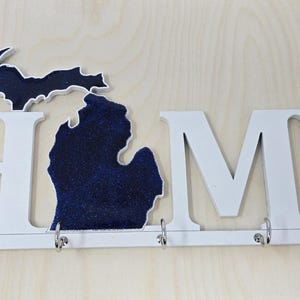 May include: White wooden "HOME" sign with a blue glitter Michigan state outline in the center. The sign has five hooks for hanging keys or other items.