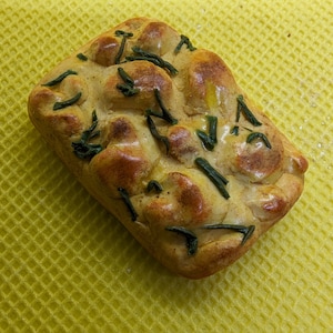 Könnte beinhalten: Ein Miniatur-Focaccia-Brot, rechteckig geformt, mit goldbrauner Kruste und dunkleren braunen Flecken. Grüne Kräuter sind über die Oberseite verstreut. Das Brot liegt auf einer gelben, strukturierten Oberfläche.