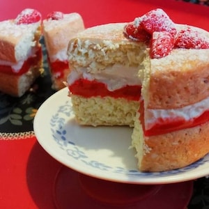 May include: A sponge cake with a slice removed, showing layers of cream and red filling. Fresh strawberries and icing sugar top the cake. Two cake slices are in the background. The cake is on a plate.
