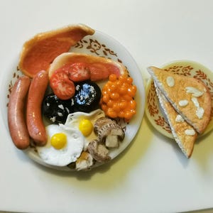 May include: A miniature full English breakfast on a white plate, including bacon, sausages, tomatoes, black pudding, baked beans, fried eggs, and mushrooms. Two slices of toast are on a separate plate. The food is arranged on a white surface.