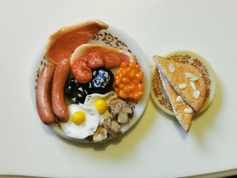 May include: A miniature full English breakfast on a white plate, including bacon, sausages, tomatoes, black pudding, baked beans, fried eggs, and mushrooms. Two slices of toast are on a separate plate. The food is arranged on a white surface.