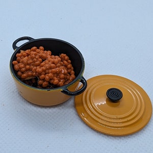 May include: A miniature orange and black enamel pot filled with baked beans, with the lid beside it. The pot has black handles and a black knob on the lid. The beans are a reddish-brown color.