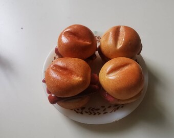 May include: Four miniature burger sliders on a white plate with a brown floral design. The buns are golden brown with a glossy finish, and the fillings are a reddish-brown color.