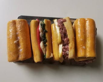 May include: Four miniature, golden-brown submarine sandwiches on a silver tray. The sandwiches are cut open, revealing colorful fillings of red, green, white, and a pinkish-brown meat. The bread has a glossy finish.