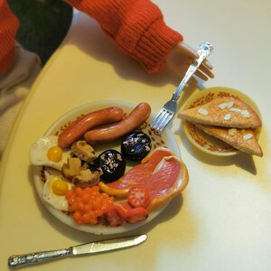 May include: Miniature full English breakfast on a white plate, including sausages, eggs, beans, mushrooms, tomatoes, and black pudding. A small plate of pizza slices is also present. A fork and knife are next to the plate. The scene is set on a light yellow surface.