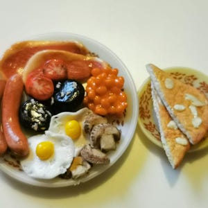 May include: A miniature full English breakfast on a white plate, featuring bacon, sausages, tomatoes, black pudding, baked beans, fried eggs, and mushrooms. Two slices of toast with almonds are on a small plate.