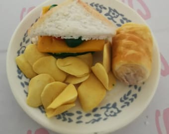 May include: A plate with a sandwich, chips, and a pastry roll. The sandwich has white bread, yellow cheese, and green and red fillings. The chips are yellow and the pastry roll is golden brown. The plate has a blue and white floral design.