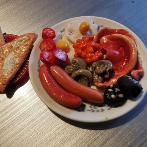 May include: A miniature breakfast plate with a variety of food items. Includes two sausages, mushrooms, tomatoes, beans, and a fried egg. Two slices of toast are on a separate red plate.