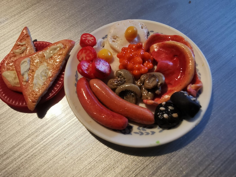 May include: A miniature breakfast plate with a variety of food items. Includes two sausages, mushrooms, tomatoes, beans, and a fried egg. Two slices of toast are on a separate red plate.