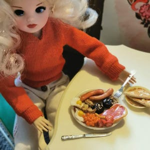 May include: A doll with blonde hair and an orange sweater sits at a table with a miniature breakfast plate. The plate includes eggs, sausage, beans, and other breakfast foods. A knife and fork are on the table.