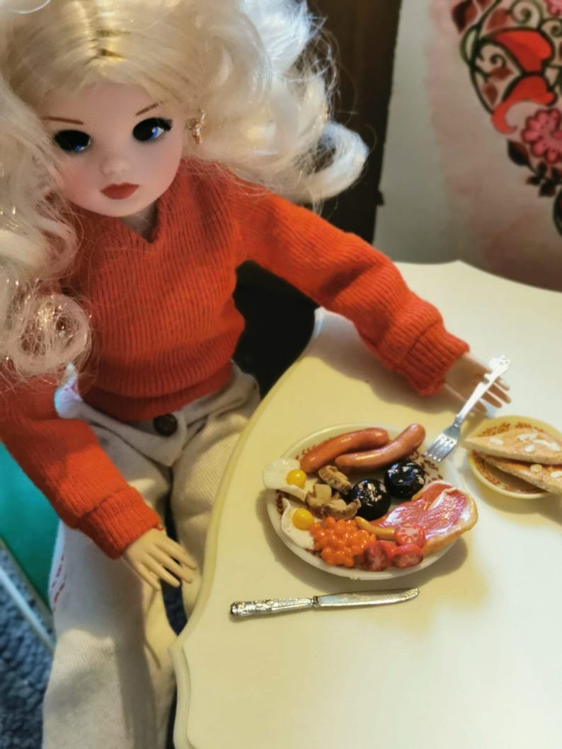 May include: A doll with blonde hair and an orange sweater sits at a table with a miniature breakfast plate. The plate includes eggs, sausage, beans, and other breakfast foods. A knife and fork are on the table.