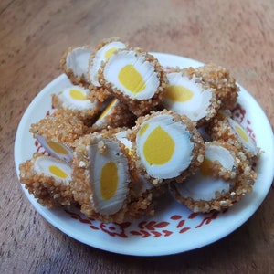 Puede incluir: Huevos empanados y fritos, cortados por la mitad para revelar la yema amarilla. Están dispuestos en un plato blanco con un diseño floral rojo. La comida está sobre una superficie de madera.