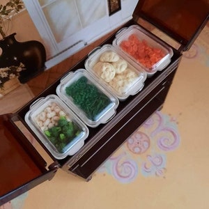 Peut inclure: Une boîte en bois brun foncé avec un couvercle à charnière, contenant quatre récipients en plastique transparent. Chaque récipient contient des éléments de couleurs différentes : blanc, vert, orange et beige. La boîte est ouverte, révélant le contenu.