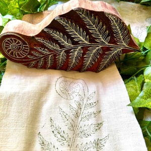 Könnte beinhalten: Ein geschnitzter Holzblock mit einem detaillierten Farnwedel- und Spiral-Fiddlehead-Design, platziert über einem hellbeigen Stoffstreifen, der einen passenden grünen Tintendruck desselben botanischen Farnmusters zeigt.