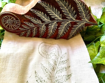 May include: A carved wooden block featuring a detailed fern frond and spiral fiddlehead design, positioned above a light beige fabric strip that displays a matching green ink print of the same botanical fern pattern.