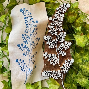 Può includere: Un timbro in legno marrone per stampa a blocchi con un design a foglia di felce intagliato e pigmento bianco nelle scanalature, posizionato accanto a un pezzo rettangolare di tessuto color avorio con una stampa a inchiostro blu.