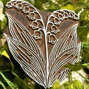 Può includere: Un blocco di stampa in legno marrone scuro con un disegno intagliato di due steli di mughetto con fiori a forma di campana e foglie lunghe e appuntite. Le linee intagliate sono profonde e definite, creando un rilievo per l'applicazione dell'inchiostro.