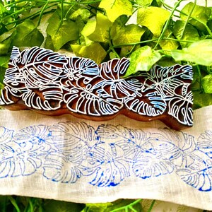 May include: A rectangular wooden block stamp featuring a carved monstera leaf pattern, positioned above a strip of light-coloured fabric displaying a repeating blue ink print of the same monstera leaf design, showcasing the block's function.