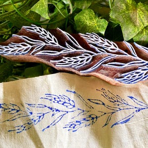 Può includere: Un blocco di legno intagliato con un motivo dettagliato di spiga di grano con residui di inchiostro blu sulle superfici in rilievo, posizionato sopra un pezzo di tessuto beige chiaro che mostra un disegno di spiga di grano blu stampato.