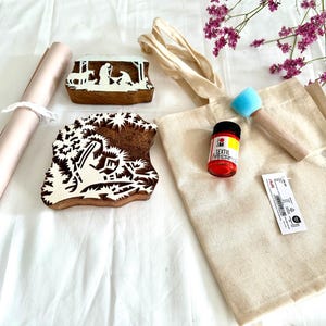 Op de afbeelding: Een knutselset met houten stempels, een canvas draagtas, rode textielverf, een schuimborstel en een rol papier. De stempels hebben gedetailleerde ontwerpen, waaronder een kerststal. De set ligt op een wit oppervlak.