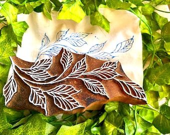May include: A brown wooden printing block featuring a carved leaf branch design with white highlights. Behind the block lies a white fabric cloth displaying a blue ink print of the same leaf branch pattern, surrounded by green artificial ivy leaves.