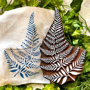 May include: A brown wooden block printing stamp featuring a carved fern leaf design, positioned next to a white fabric cloth displaying a blue ink-stamped fern leaf print. The stamp surface shows the raised white-painted fern pattern clearly.